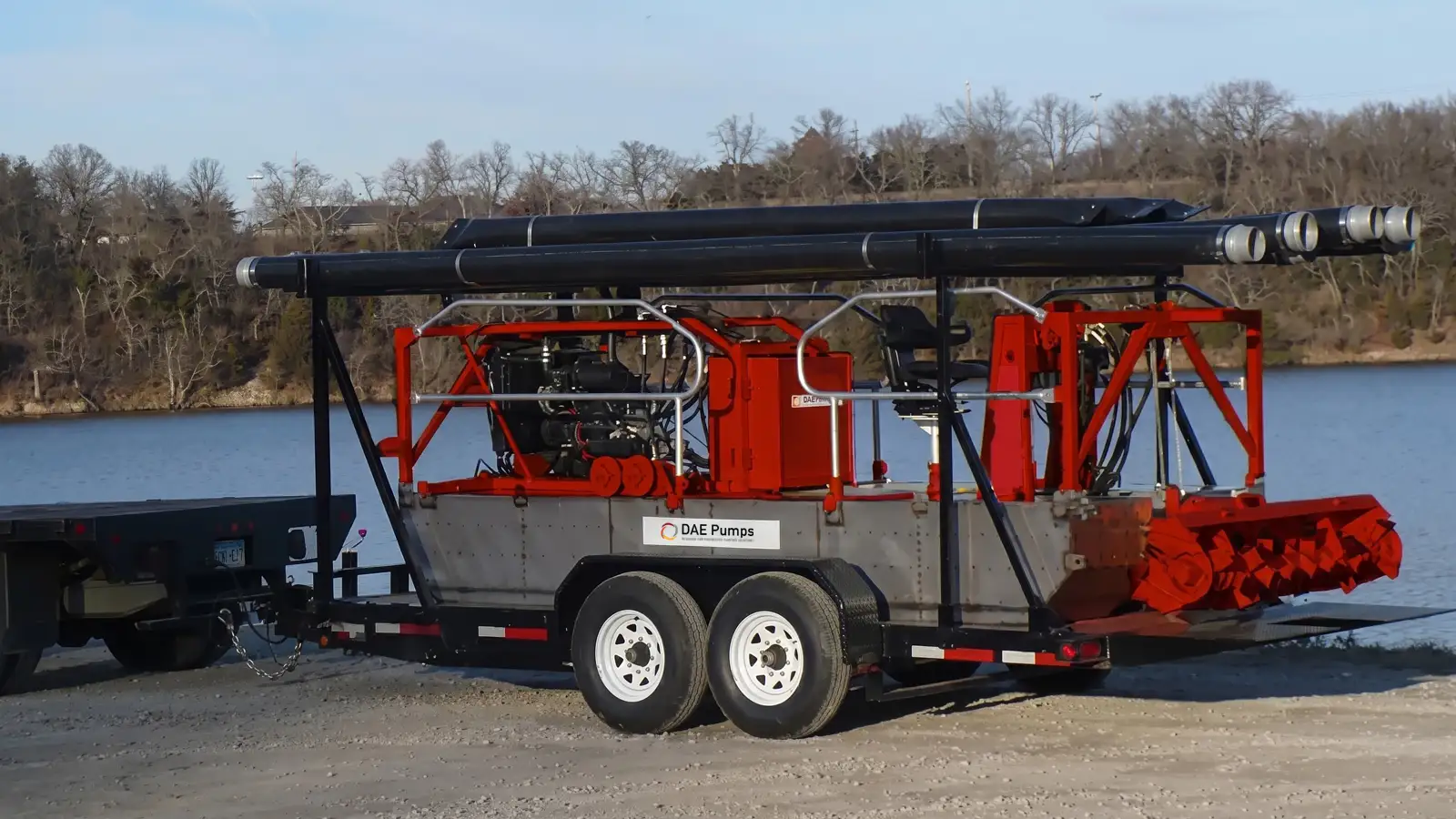 Compact, Trailerable Pond Dredge Sediment Slayer pond dredge loaded on a tandem-axle trailer beside a lake