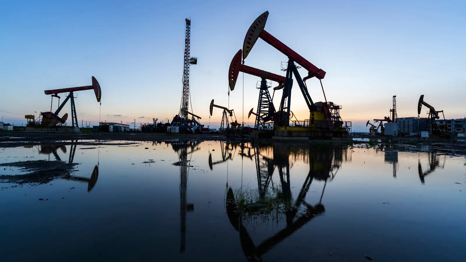 Oil and Gas Pumping Oil pumpjacks and a drilling rig reflected in water at sunset